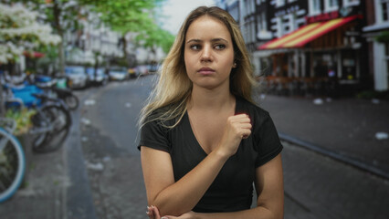 Woman with fist to chest on a city street, wearing black v neck tee and crossing one arm across waist; concerned reflection.