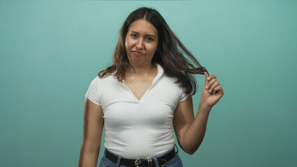 Woman in white shirt twirling her hair with right hand and pouting while posing in a teal studio;...