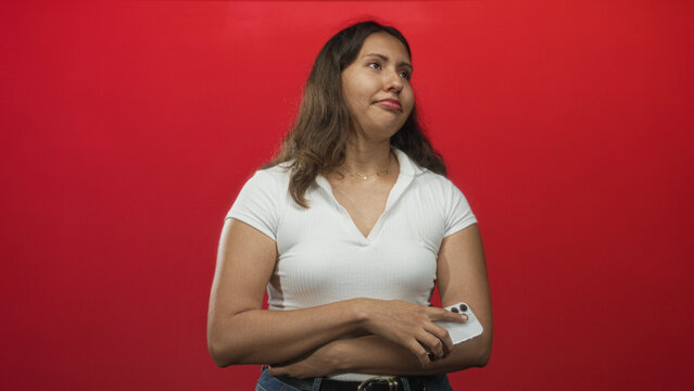 Young latina woman holding smartphone and looking down in a red studio, wearing white top and belt; frustration. - Powered by Adobe