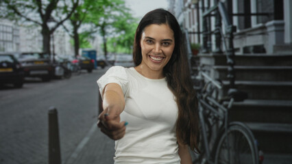 Fototapeta premium Woman extends hand in handshake gesture toward camera on urban street lined with bicycles and parked cars; friendliness.
