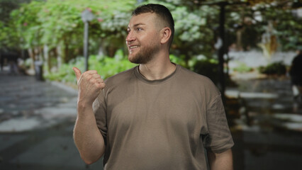 Fototapeta premium Man smiling and giving thumbs up with bare thumb and hand visible on a leafy street path beside green park trees, wearing casual tshirt; approval.