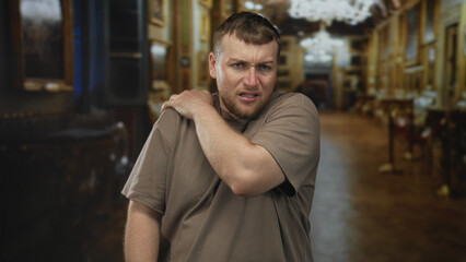 Obraz premium Young heavyset man holding shoulder, grimacing with forearm exposed in museum gallery corridor, short beard visible; discomfort tension.