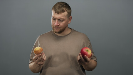 Man holding red apple and muffin with visible hands in gray studio backdrop, weighing food choice...