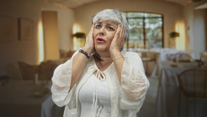 Woman holding hands to face and looking upward in a restaurant building dining room with bare forearms visible; anxiety vulnerability.