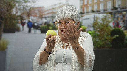 Woman holding green apple and gesturing with hand on street, smiling and pointing to chest;...