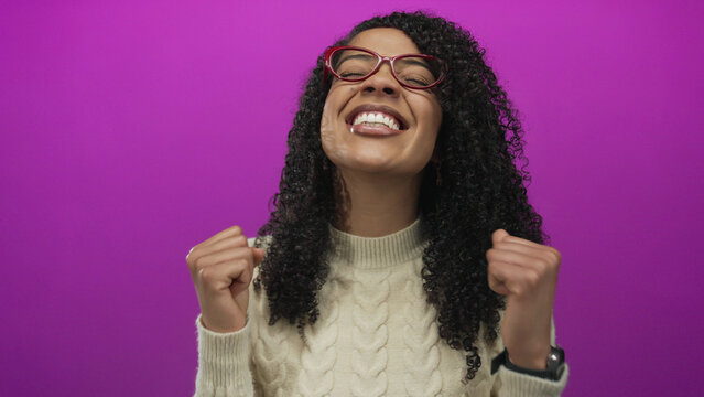 Woman wearing red glasses smiles and clenches fist in studio against purple wall; joy triumph success celebration victory happiness excitement elation euphoria bliss.