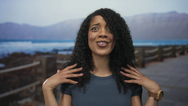 Woman flips hair with expressive hand gesture on seaside street under soft morning light; confusion. - Powered by Adobe