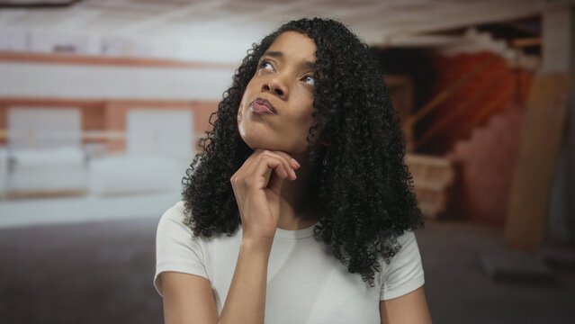 Woman with curly hair resting hand on chin inside unfinished building with exposed brick walls and scattered lumber; contemplation.