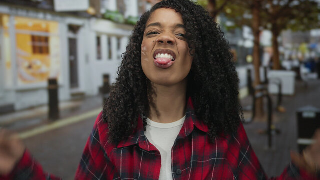 Woman with curly hair sticks out her tongue on a vibrant city street with blurred storefronts; cheeky mischief.