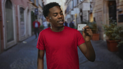 Man grips creditcard with right hand on busy urban street outdoors next to historic building facade; confidence.