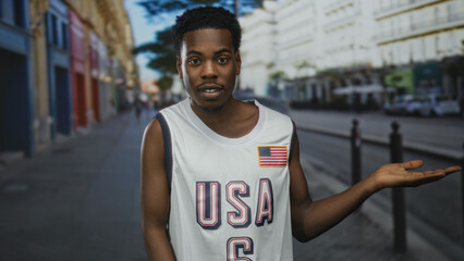African american man in sleeveless usa basketball jersey extends palm up hand to the side on city street; openness.
