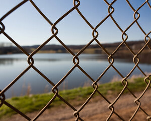 Fototapeta premium Peaceful lakeside landscape seen through rustic chain link fence, evoking a sense of security and natural beauty with reflections and serene blue sky