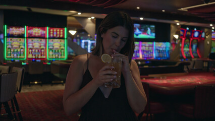 Woman holding cocktail glass, sipping through a straw and smiling, bare shoulders visible in casino; celebration night out.