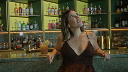 Woman smiling with bare shoulders and cocktail on marble bar counter, looking up while resting hands on edge; relaxed confidence.