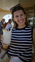 Woman holding a cold pineapple cocktail glass and sipping through a straw at a building bar table, smiling; casual summer relaxation.