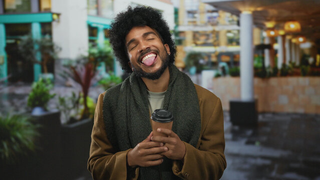 Man enjoying coffee on a bustling city street, flashing a cheerful smile amid urban surroundings, showcasing style and comfort outdoors.