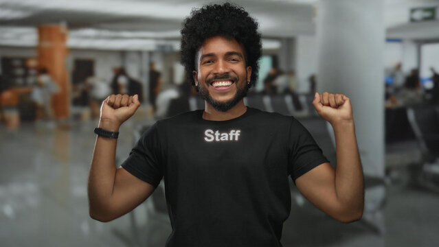 Man in black staff uniform smiling indoors at a busy airport, embodying friendliness and professionalism in a diverse, vibrant setting.