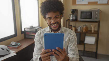 African american man smiling in an office, holding a tablet, surrounded by office essentials like a calculator and documents, conveying a work environment.