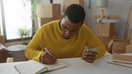 Man writing notes with smartphone in studio stacked with moving boxes and notebooks; concentration productivity.