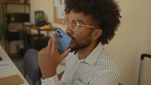 Man speaking into smartphone in modern office wearing glasses and striped shirt highlighting technology usage among professionals in indoor workplace setting with focus on communication.