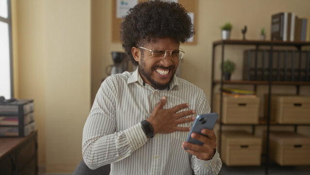 Man in glasses holding chest and phone indoors shows discomfort in an office setting, highlighting his reaction to possible chest pain.