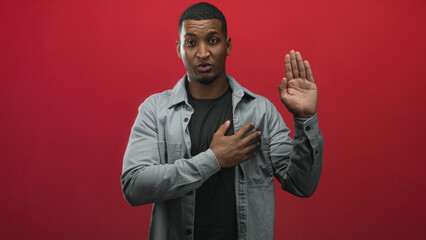 Young black man with hand on chest and raised palm making an oath gesture in studio against red...