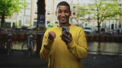 Young man holding smartphone, smiling and glancing down while wearing a yellow sweater on a street by a canal; digital connection joy.
