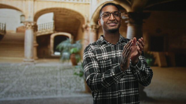 Young black man smiling and clapping hands while wearing glasses and a plaid flannel shirt in a historic building courtyard with arches and potted plant; confidence warmth. - Powered by Adobe