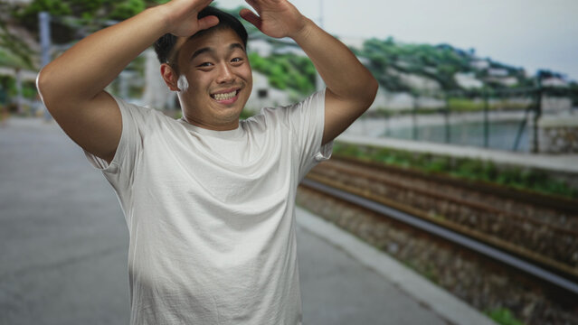 Young chinese man raises both hands beside railway track at a building, smiling confidently; optimism.