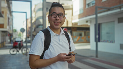 Man holding smartphone and texting on city street sidewalk with backpack and glasses; urban commute focused.