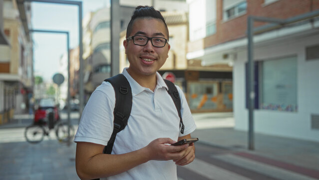 Man holding smartphone and texting on city street sidewalk with backpack and glasses; urban commute focused.