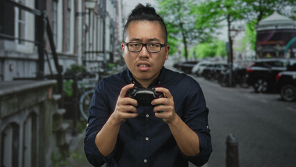 Young chinese man gripping a game controller with tense hands on a busy street; competitive frustration.