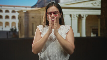 Woman smiling with hands pressed together wearing red glasses and white sleeveless top in front of building facade  gratitude. © Krakenimages.com