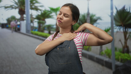 Fototapeta premium Woman rubbing shoulder with hand and wincing on a street promenade wearing denim apron and pink striped shirt; discomfort.