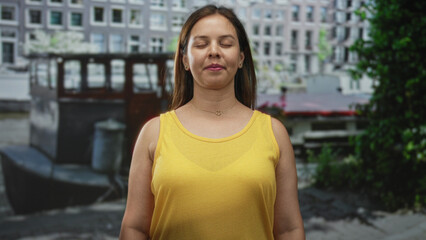 Woman with eyes closed and bare shoulders in yellow tank top standing on street by canal and buildings in amsterdam; serenity meditation.