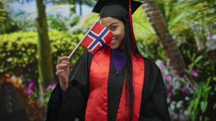 Young latin woman in cap and gown smiling and holding norway flag amid lush forest; pride achievement.