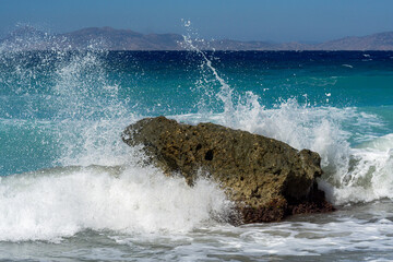 Powerful turquoise sea waves crashing against a lone coastal rock, sending white spray into the air on a sunny day with distant mountains on the horizon.