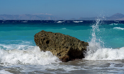 Powerful turquoise sea waves crashing against a lone coastal rock, sending white spray into the air on a sunny day with distant mountains on the horizon.