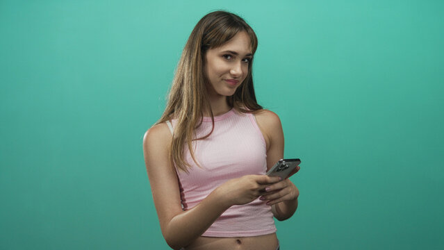 Young woman wearing pink tanktop holding smartphone and texting while looking down in a teal studio with plain colored wall; thoughtful.