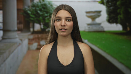 Girl with closed eyes and bare shoulders standing in a green courtyard by a stone fountain and building; inner peace reflection tranquility.