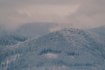 A hazy, layered view of snow-dusted mountains and valleys, with low-hanging clouds obscuring the peaks, creating a deep sense of mystery, scale, and quiet winter atmosphere.