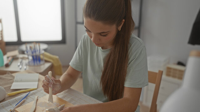 Teenage girl sculpts white clay on wooden board in studio under daylight; creativity craftsmanship concentration. - Powered by Adobe