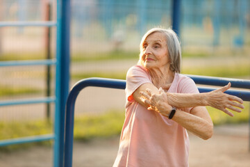 A happy, active elderly woman in a pink shirt stretches her arms outdoors near blue gym equipment. Her smile and focus highlight senior fitness, health, and an active lifestyle.