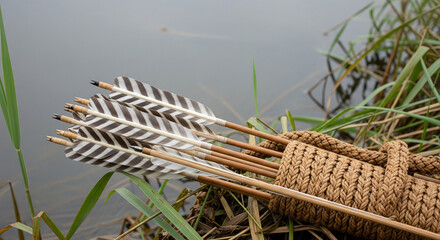 A close-up of a woven or braided quiver holding a bundle of traditional arrows with natural brown and white striped feather fletchings, leaning against tall reeds