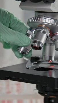Vertical close-up shot of unrecognizable gloved researcher operating microscope while carrying out experiment in laboratory