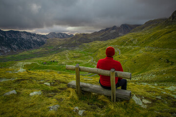A person in a red jacket sits on a wooden bench overlooking a dramatic mountain landscape under dark, moody clouds.