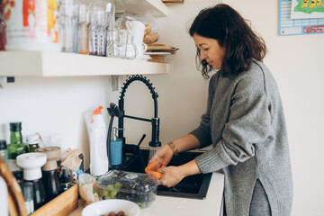 Woman with dark hair wearing a gray sweater is washing vegetables at a modern kitchen sink, surrounded by various kitchenware and a bright, inviting atmosphere