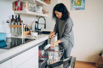 Asian woman in gray sweater unloading clean dishes from a modern dishwasher in a stylish kitchen,...