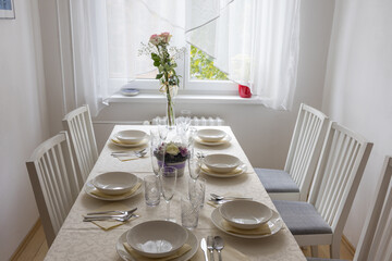 Set dining table for lunch by a window, in a modern interior. The shot captures a dining table with a white tablecloth, perfectly set for six people in a modern interior featuring white walls