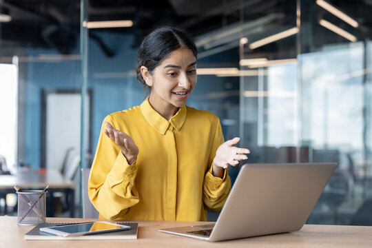 Smiling young businesswoman sitting at a desk in the office and talking on a video call on a laptop, gesturing with her hands